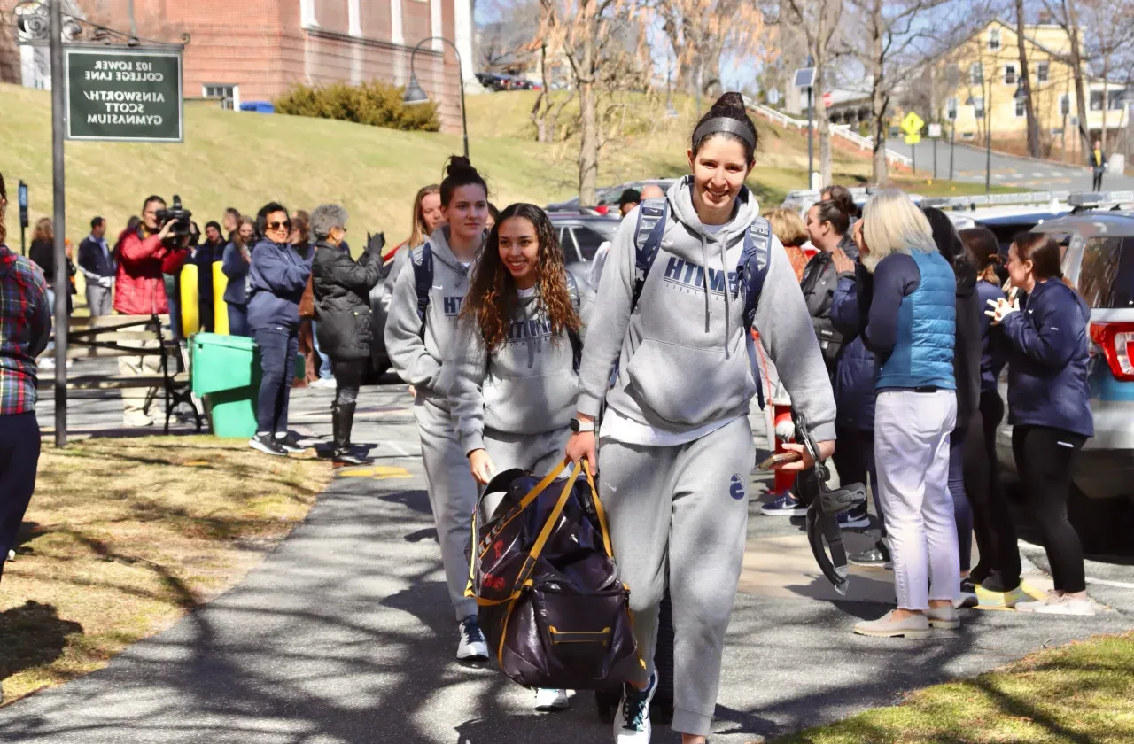 Basketball players, carrying luggage and smiling, walk along the path toward the bus